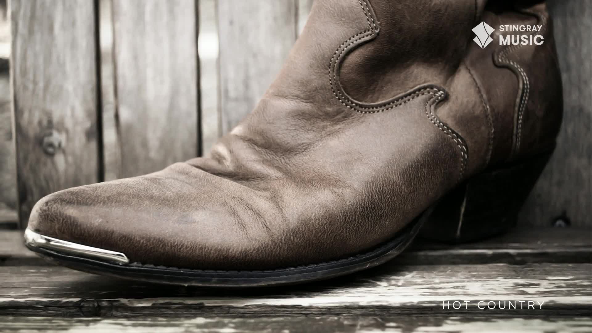 A worn leather cowboy boot rests on weathered wooden planks. The toe cap gleams with a silver accent, hinting at a Canadian country tune.