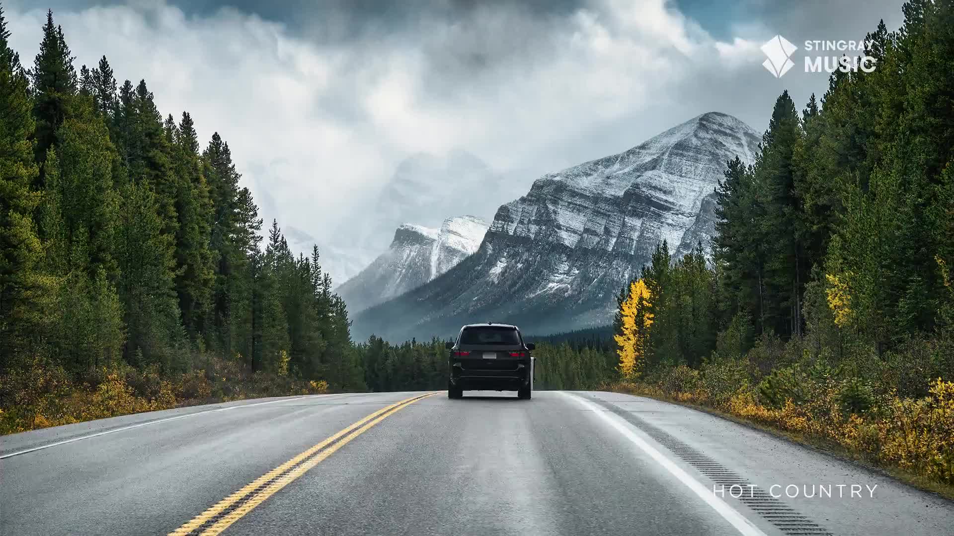 A dark SUV drives down a highway in the Canadian Rockies. Snow-capped mountains rise behind a dense forest of evergreens.