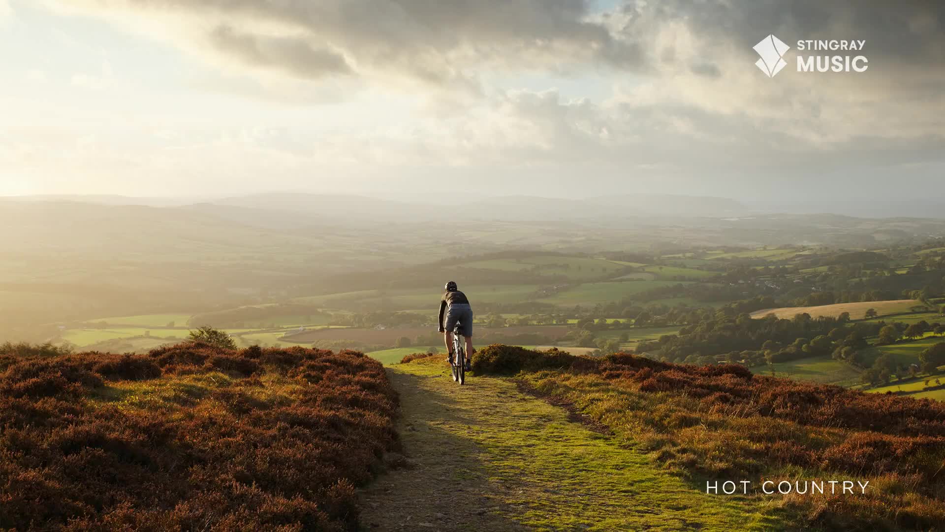 A lone cyclist pedals along a dirt path, the vast Canadian landscape unfolding before them. The sun casts a warm glow over rolling hills and distant fields, creating a peaceful, expansive scene.