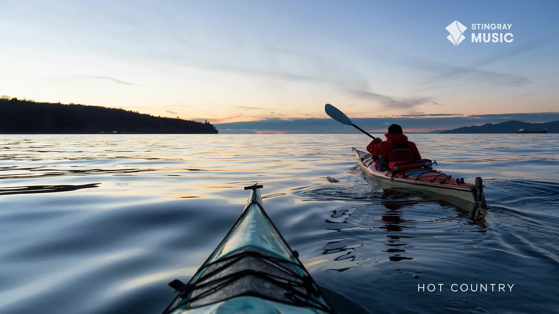 A kayaker in a bright orange jacket paddles through calm water as the sun sets over a forested coastline in British Columbia. Another kayak, viewed from the front, cuts through the gentle waves ahead.