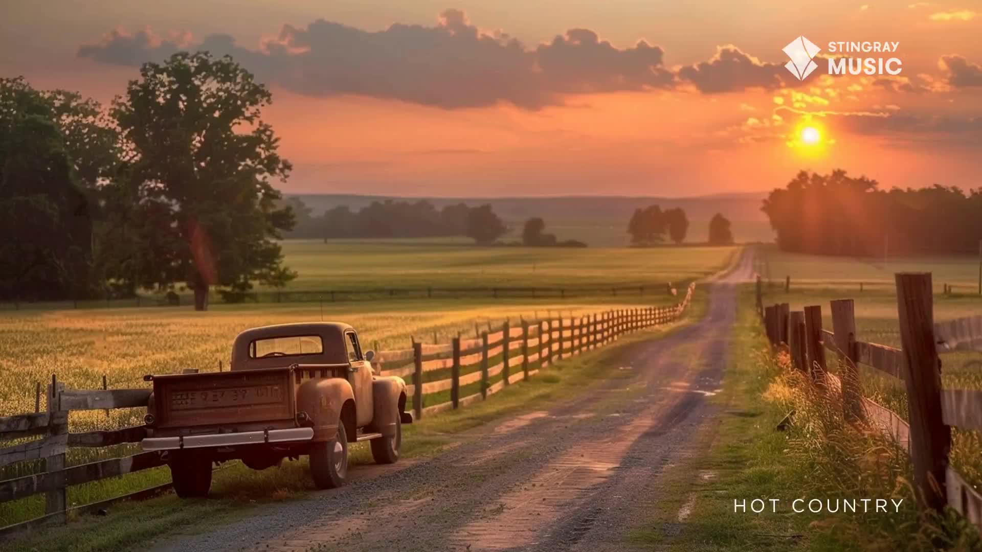 The sun dips low, painting the sky in fiery hues over a dusty Canadian road. An old pickup truck sits parked beside a weathered wooden fence, its bed empty.