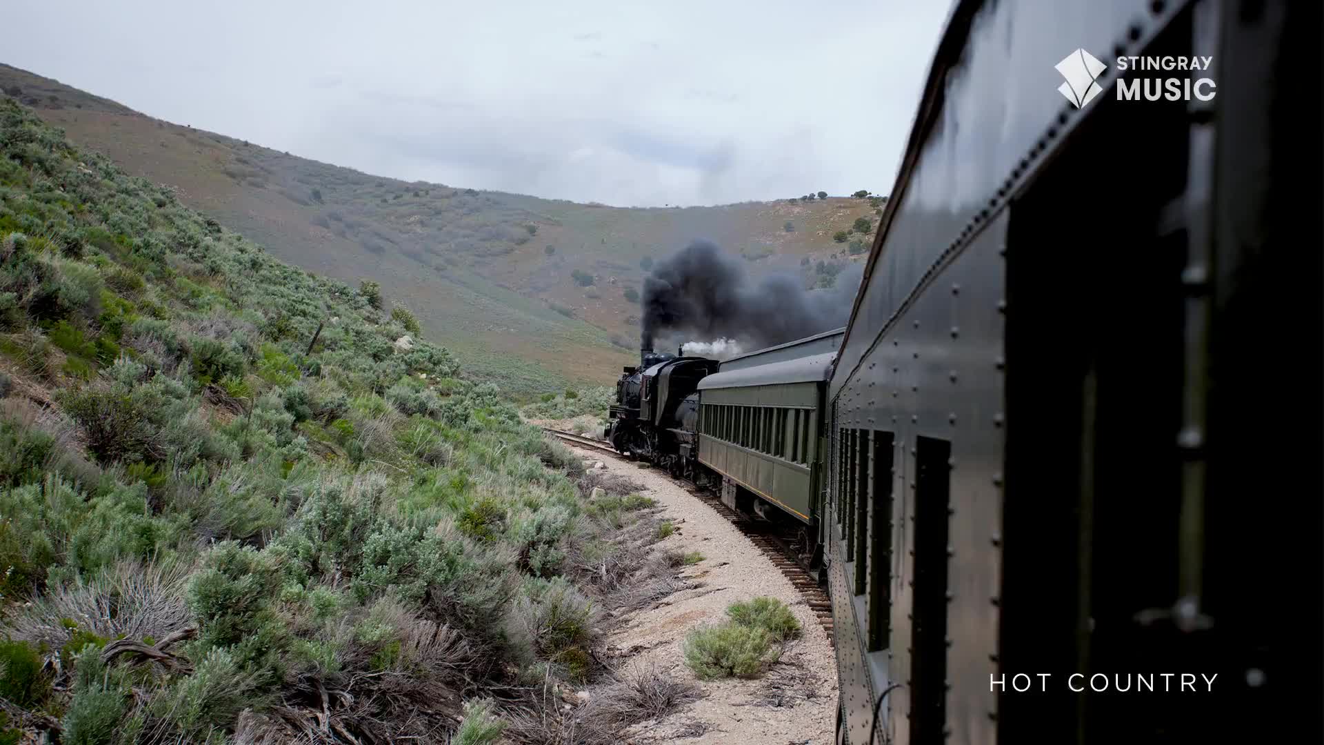 A steam locomotive pulls a string of green passenger cars around a bend, its black smoke billowing against the grey sky. The train winds through a rugged, sagebrush-covered hillside in what feels like the Canadian wilderness.