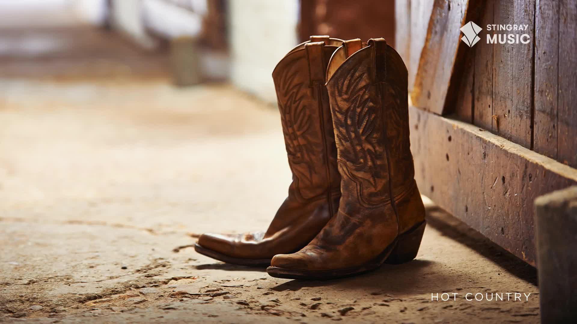 A pair of well-worn leather cowboy boots stands ready by a rustic wooden door. The warm, dusty light suggests a quiet moment before heading out to enjoy some hot country music.