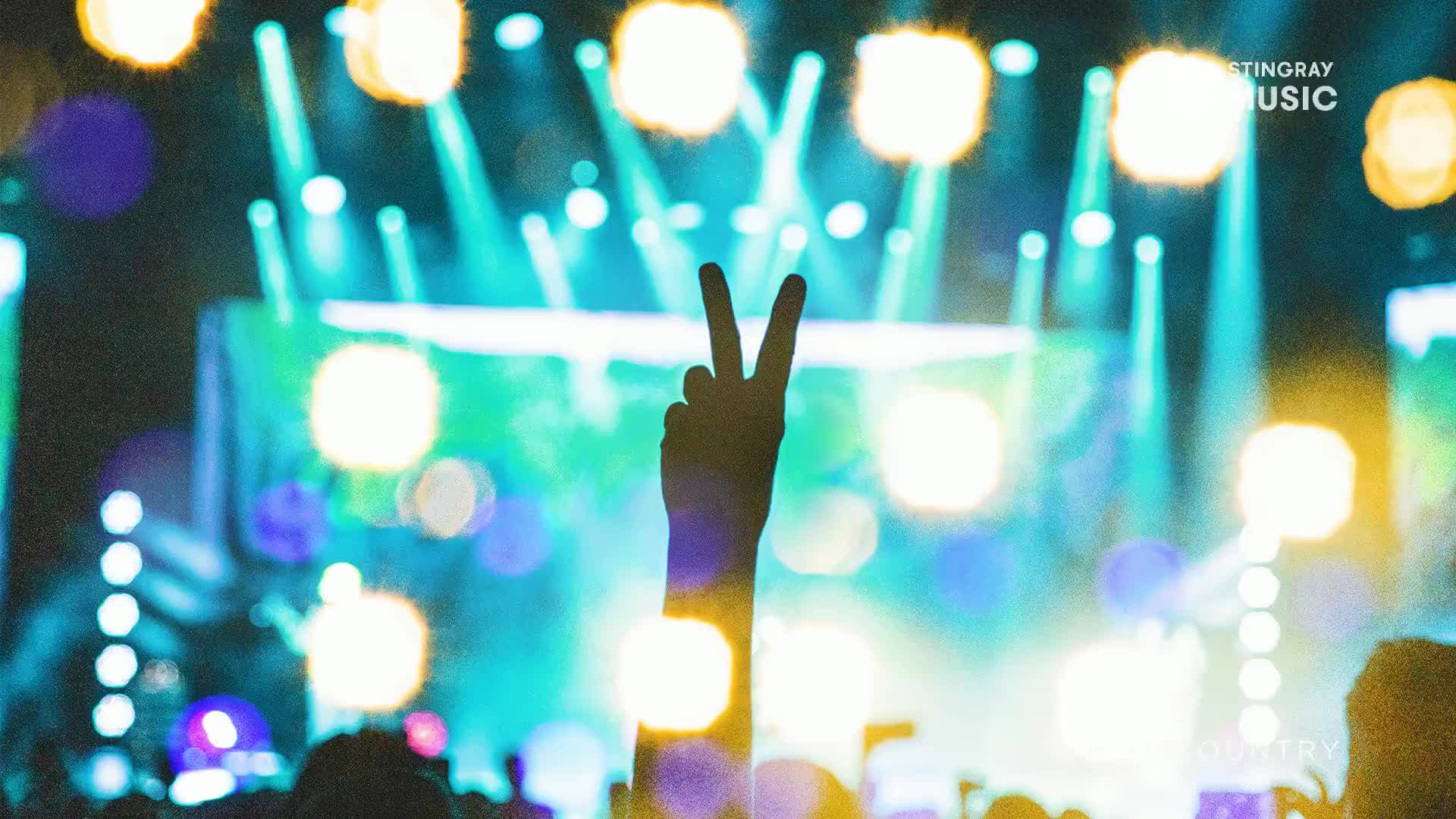 A hand holds up two fingers against a backdrop of bright stage lights. The crowd is packed together, a sea of silhouettes enjoying the Stingray Hot Country performance.