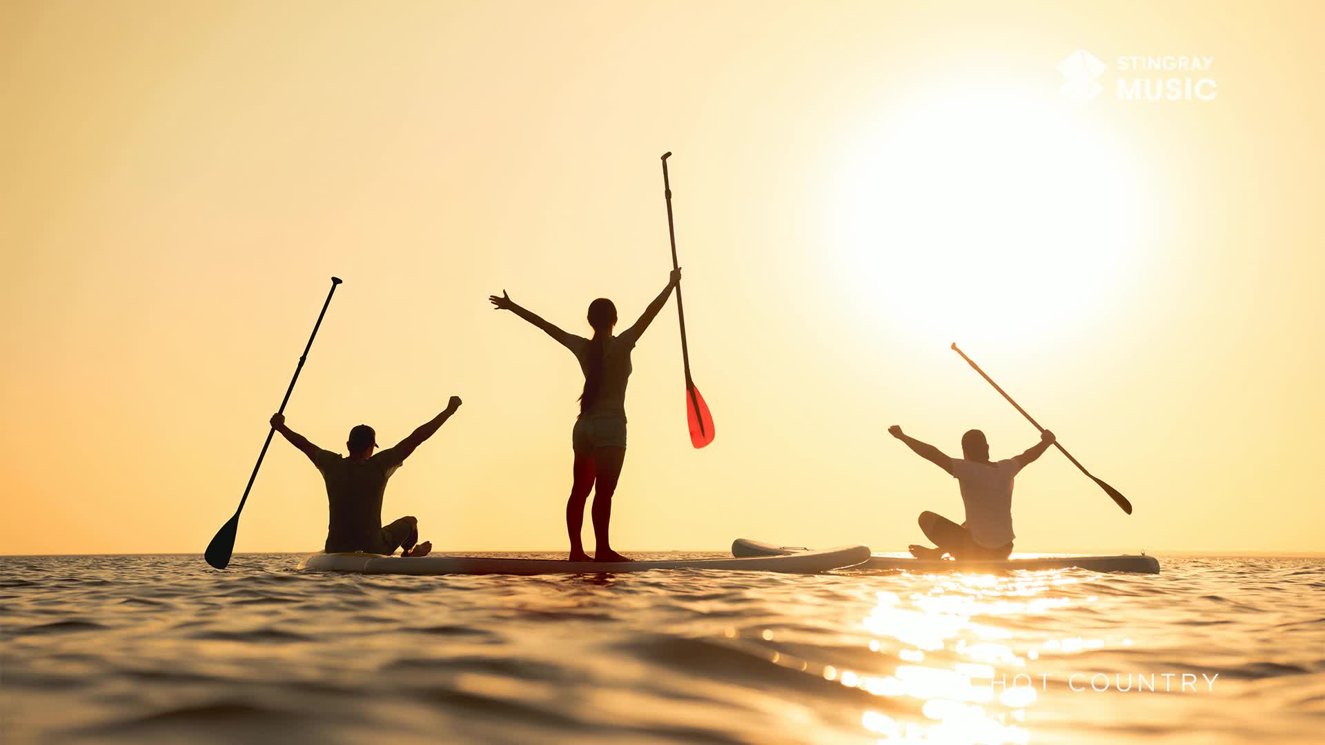 Three people are paddleboarding on calm water as the sun sets over the horizon. Their silhouettes are stark against the bright sky, each with arms raised in celebration.