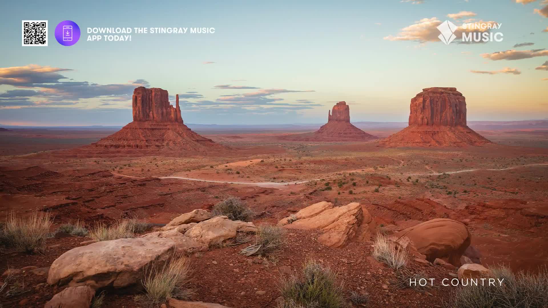The sun dips low, painting the vast desert landscape in warm hues. Three massive sandstone buttes stand sentinel against the fading light, their red rock formations stark against the pale sky.