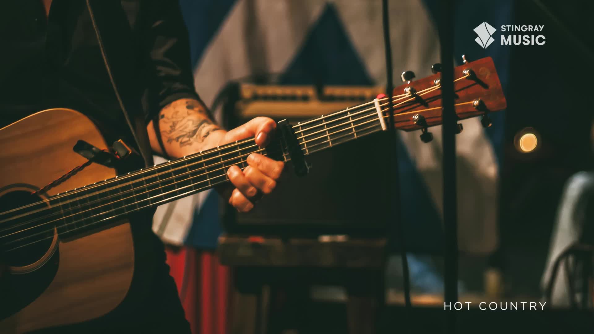 Fingers dance across the fretboard of an acoustic guitar, a capo clamped near the nut. The warm glow of stage lights catches the wood grain as a country song plays.