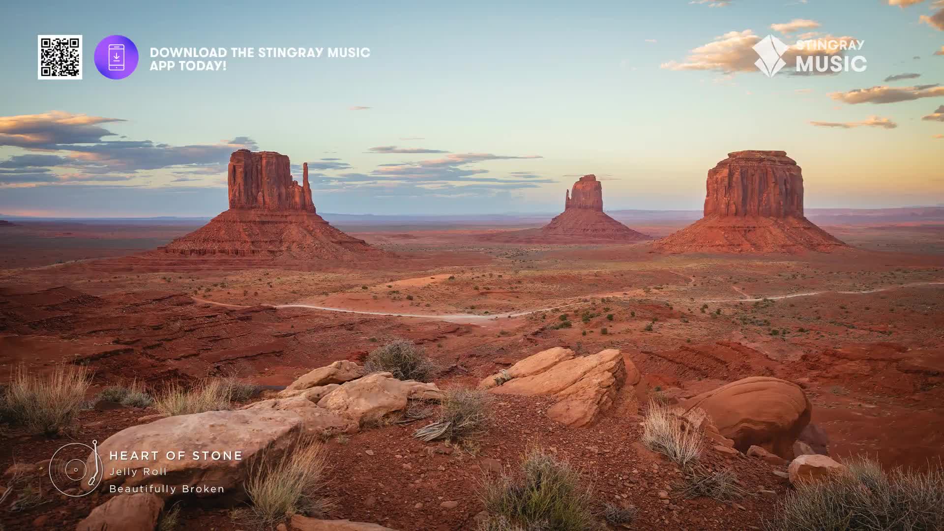 The sun dips behind massive red rock formations, casting long shadows across the desert floor. A dirt road winds through the vast landscape, leading towards the iconic mesas.