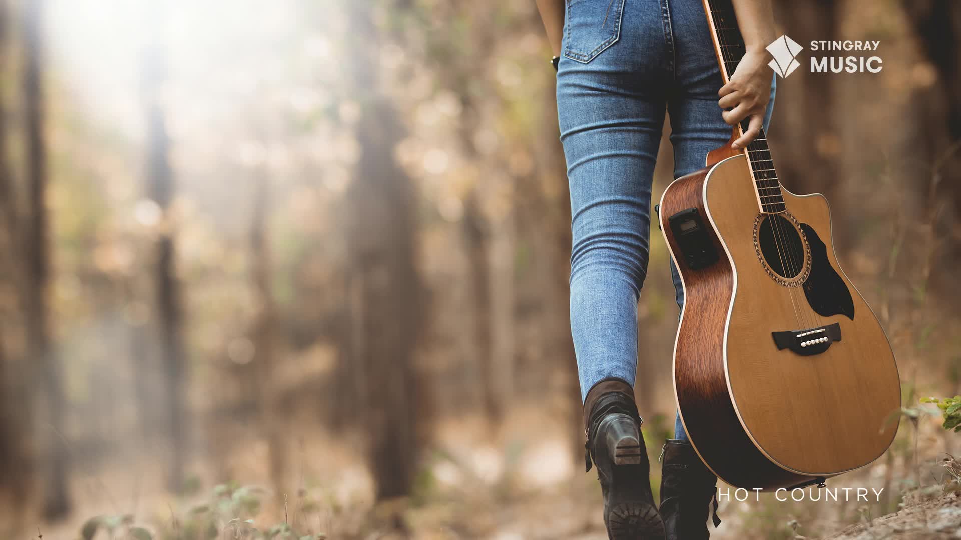 A person walks through a sun-dappled forest, carrying an acoustic guitar. The denim-clad legs and worn boots suggest a journey, perhaps to a gig or just a quiet moment with music.
