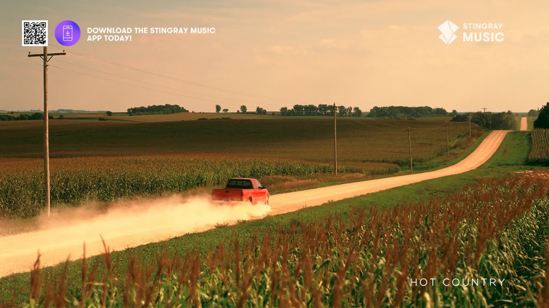 A red pickup truck kicks up a plume of dust as it speeds down a gravel road through Canadian farmland. The truck's tires churn the dirt, creating a hazy trail that hangs in the warm, late afternoon air.