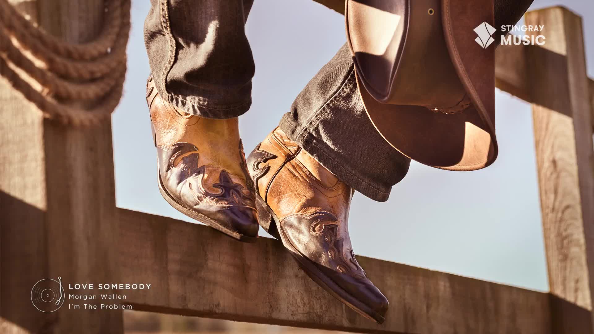 Cowboy boots rest on a wooden fence, the sun catching the intricate stitching. A wide-brimmed hat hangs nearby, hinting at a day spent outdoors in the Canadian countryside.