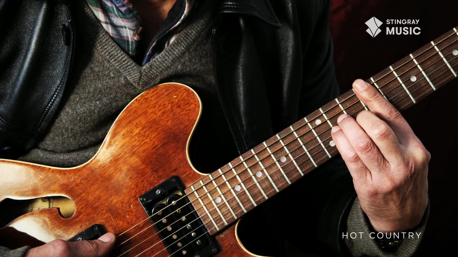 A man's hands are on a sunburst electric guitar, fingers pressing down on the fretboard. The warm wood grain of the instrument catches the light as he prepares to play.