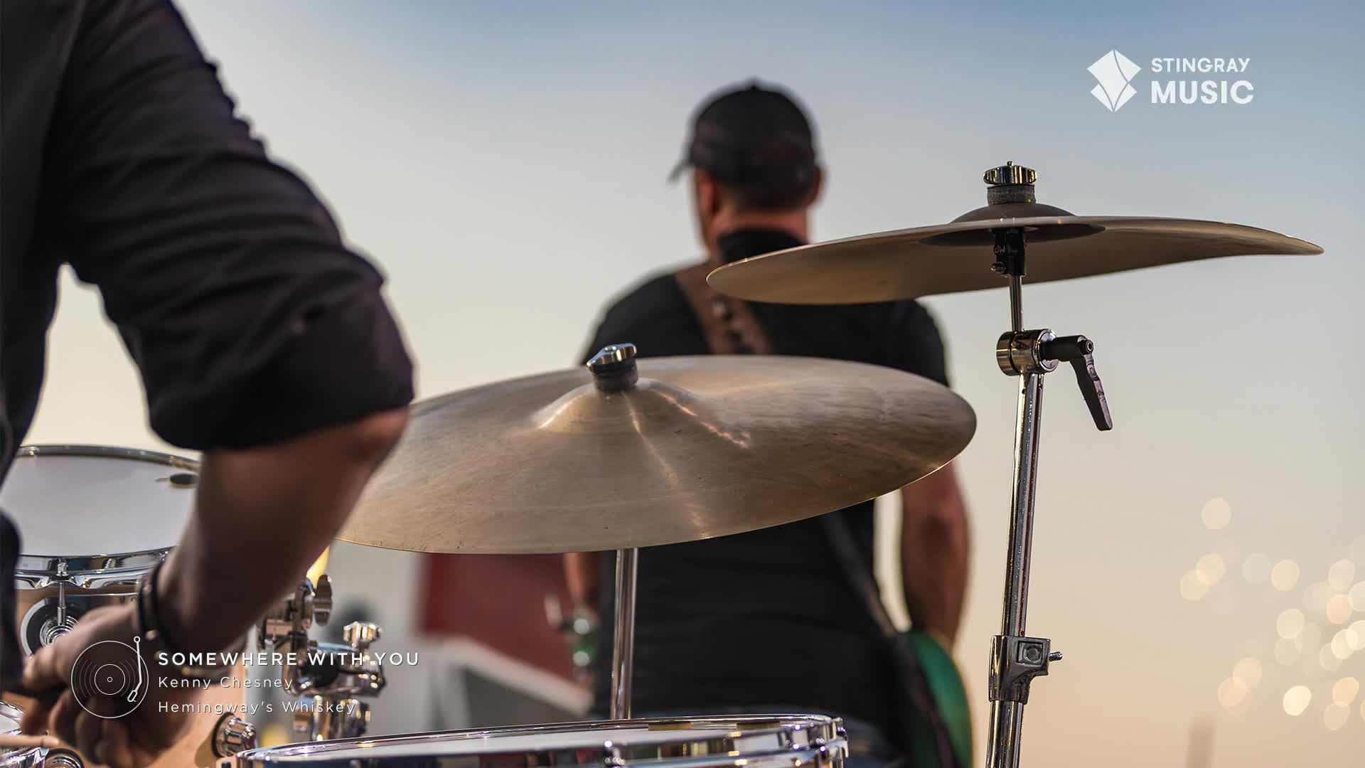 The drummer's arm is raised, poised to strike the cymbal. A band member in a black t-shirt and baseball cap stands behind the drum kit, facing away.