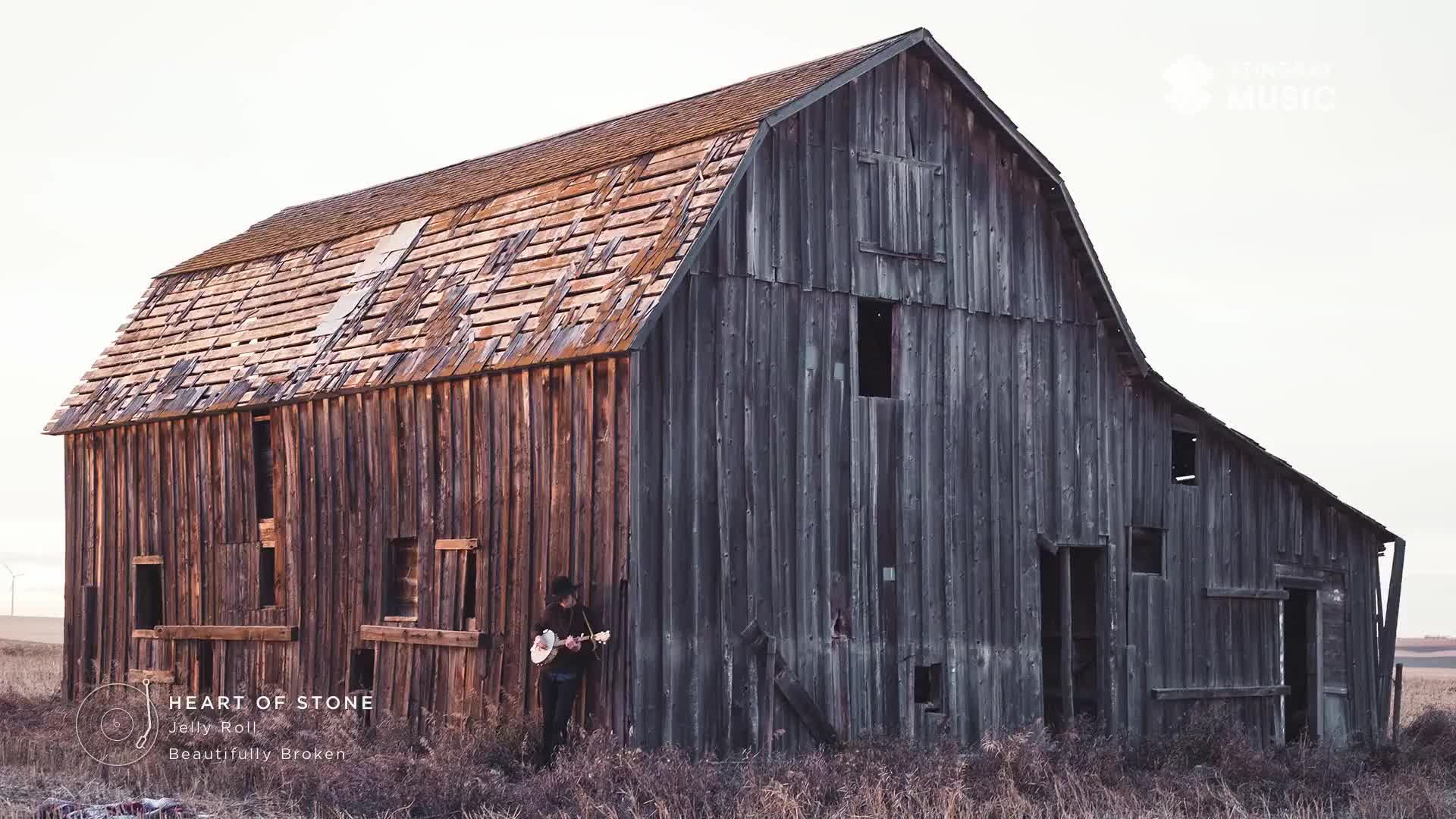 A lone figure plays an acoustic guitar outside a weathered, wooden barn in the Canadian prairie. The sky is a pale, hazy white, and dry grass surrounds the structure.