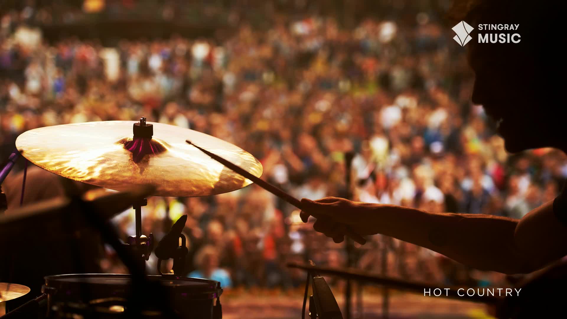 The drummer's stick strikes the cymbal, a bright flash against the shadowy silhouette. A massive crowd stretches out behind the stage, a sea of faces bathed in warm light.