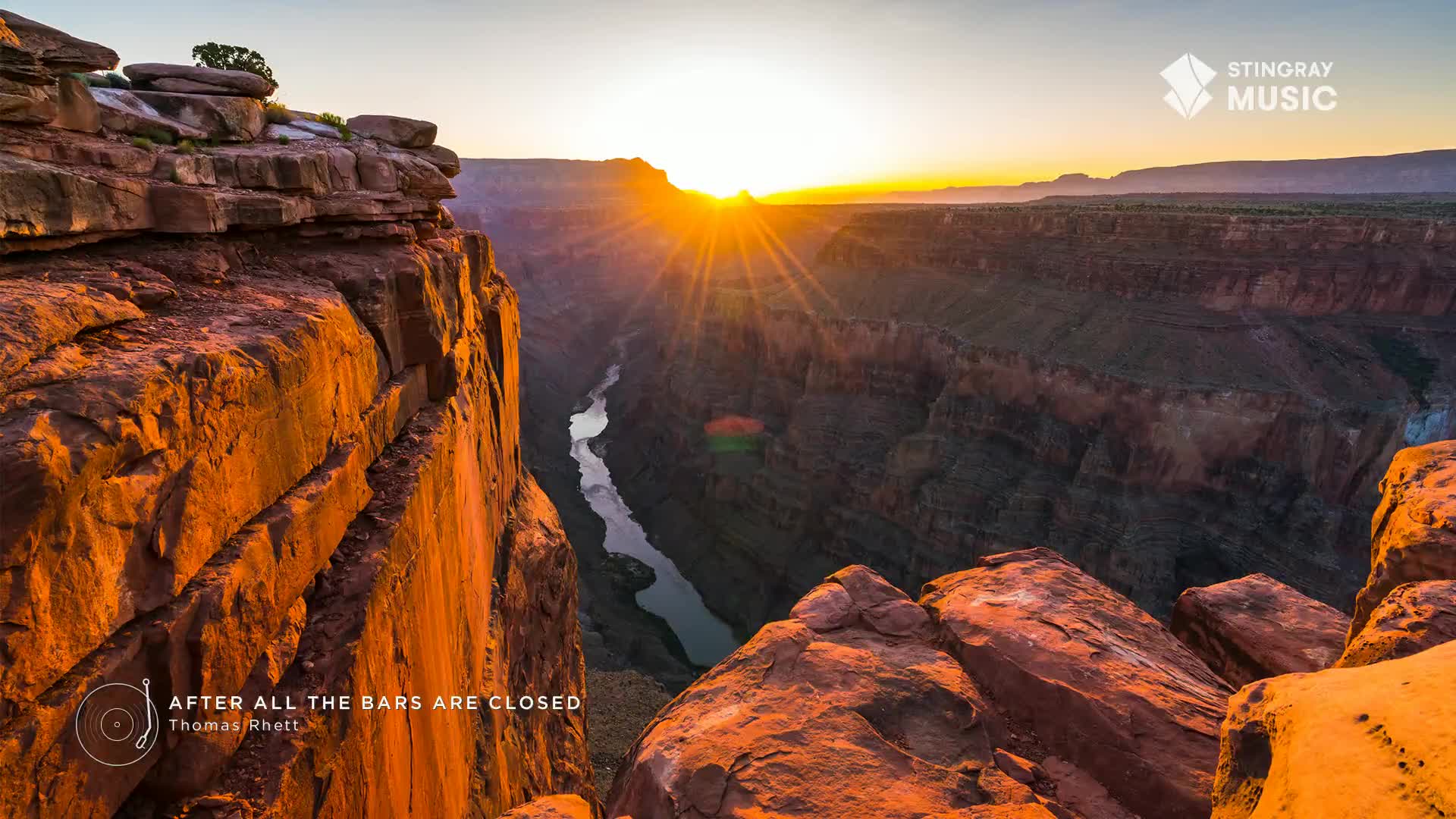 The sun dips below the horizon, casting long shadows across the vast canyon. A river snakes through the valley floor, reflecting the warm, golden light.