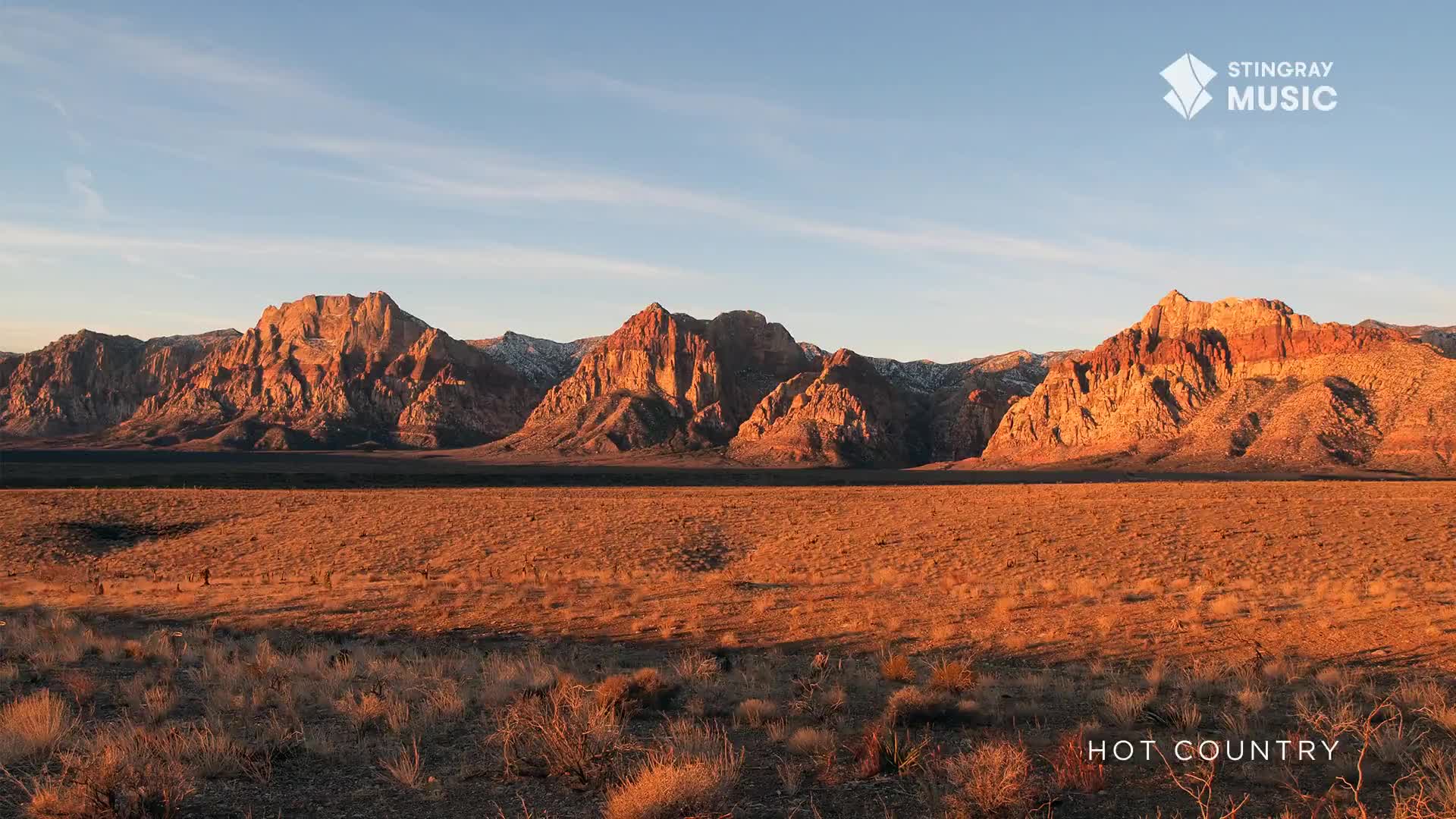 The sun casts a warm glow on the rugged, orange mountains. Dry, golden grass stretches out before them, dotted with sparse desert shrubs.