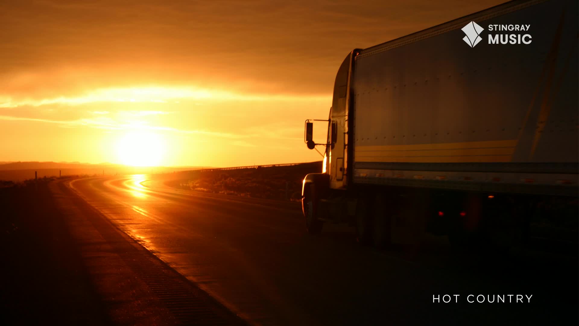 A semi-trailer truck rumbles down a highway towards a fiery Canadian sunset. The sun's brilliant rays reflect off the wet asphalt, creating a shimmering path.