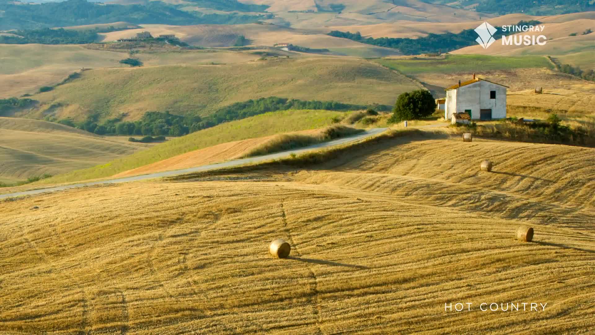 The sun casts long shadows across the golden fields, highlighting the freshly cut stubble and round bales of hay. A lone farmhouse sits atop a gentle rise, overlooking the rolling landscape.