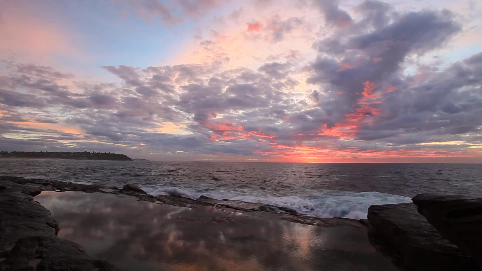 The sky is streaked with pink and orange, the colors reflected in the still water of the tide pools. Waves crash against the rocks, their white foam contrasting with the dark ocean.
