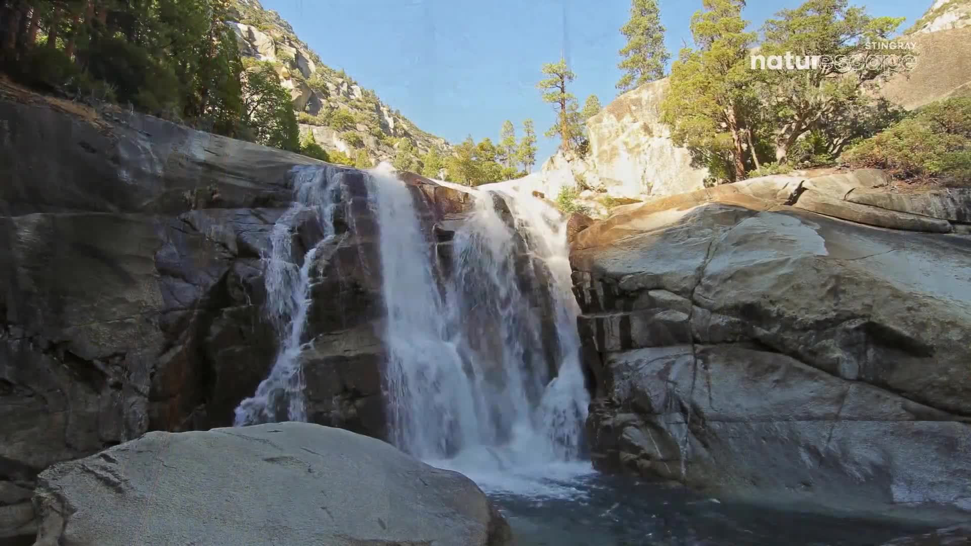 Water cascades down a series of rocky steps, forming a wide waterfall. The water's white against the dark stone, and the sun shines on the surrounding trees.
