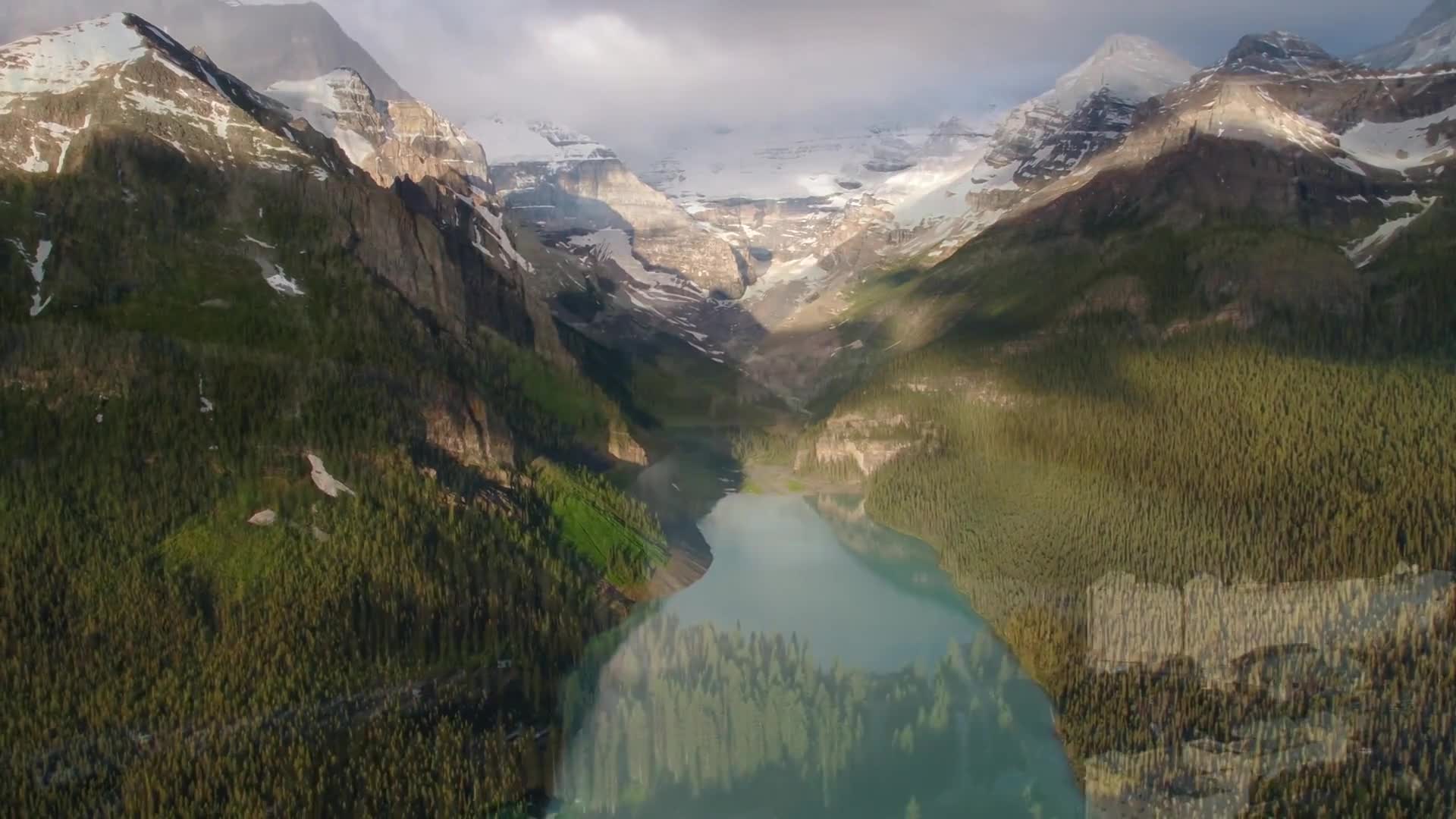 Sunlight streams through the clouds, illuminating the snow-capped peaks and casting shadows across the dense evergreen forests. A still, turquoise lake reflects the surrounding mountains, nestled in a valley in Canada.
