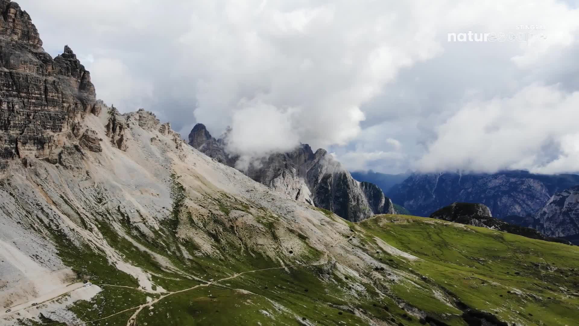 The peaks of the mountains are partially obscured by thick, low-hanging clouds. Below, the green slopes of the hills meet the barren, rocky mountainsides.
