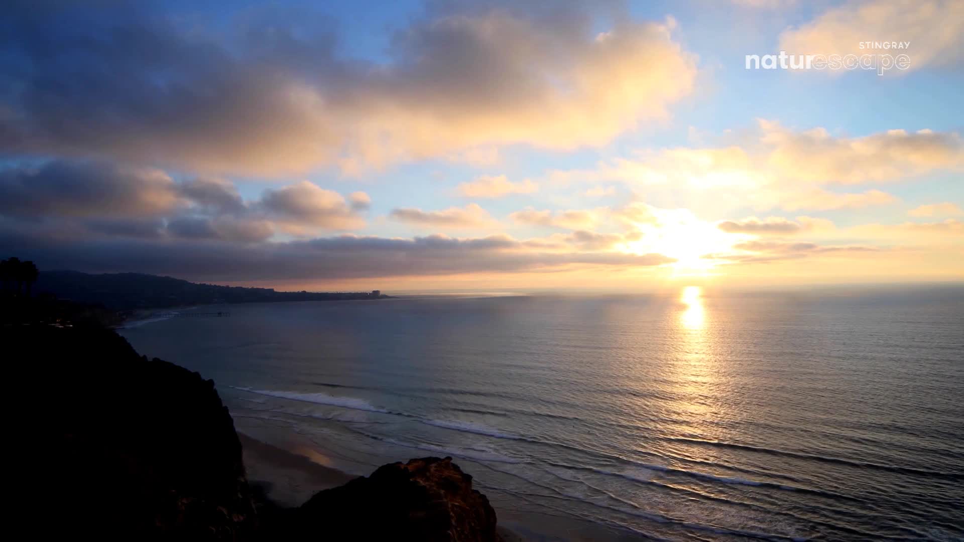 The sun, a bright orb, is sinking toward the horizon, its light reflecting on the water. Dark clouds cast shadows over the coastline, while the sky above is painted with soft hues of orange and blue.
