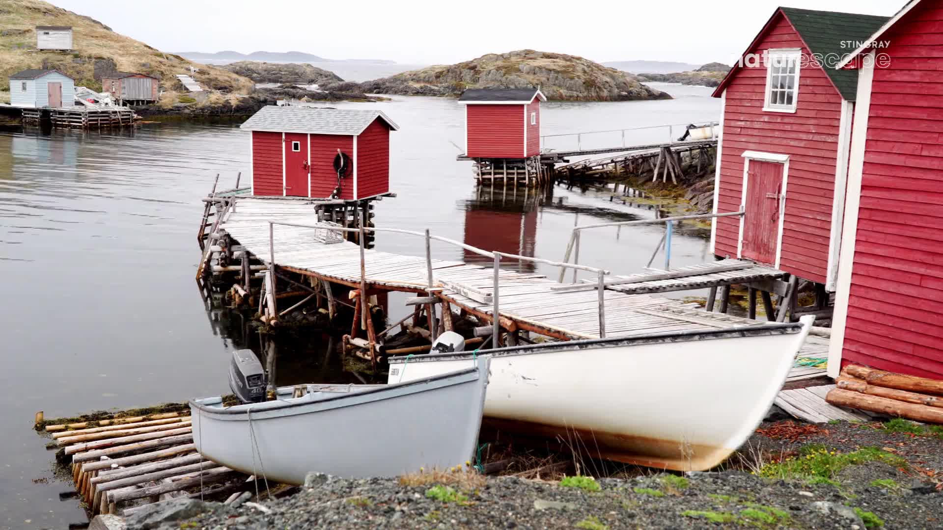 The weathered wooden docks extend into the calm water, leading to small, red fishing sheds. Two boats rest on the shore, their hulls reflecting the overcast sky.
