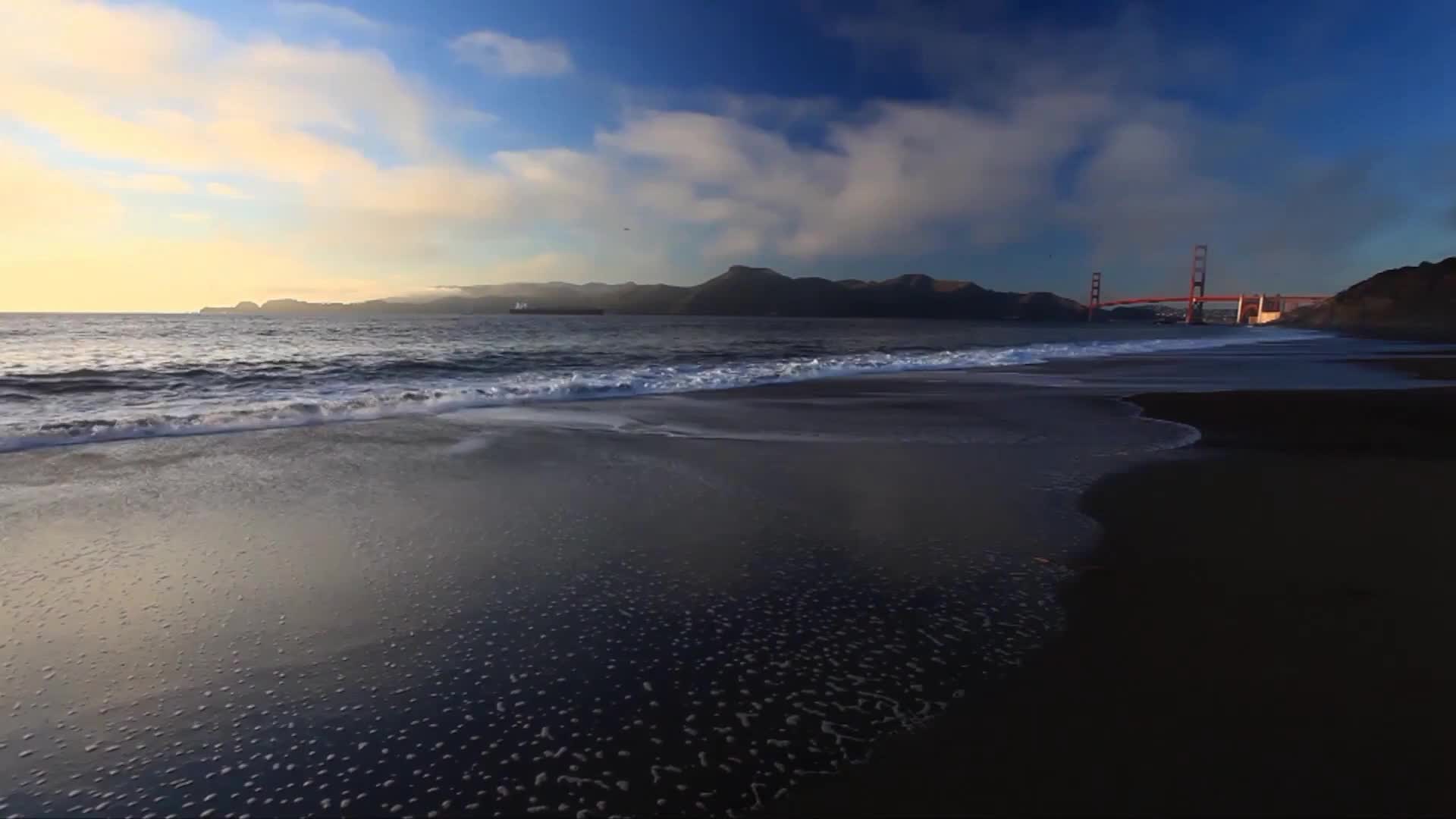 The waves roll in, leaving a foamy white fringe on the dark sand. In the distance, the Golden Gate Bridge stands silhouetted against a sky of soft blues and golds.
