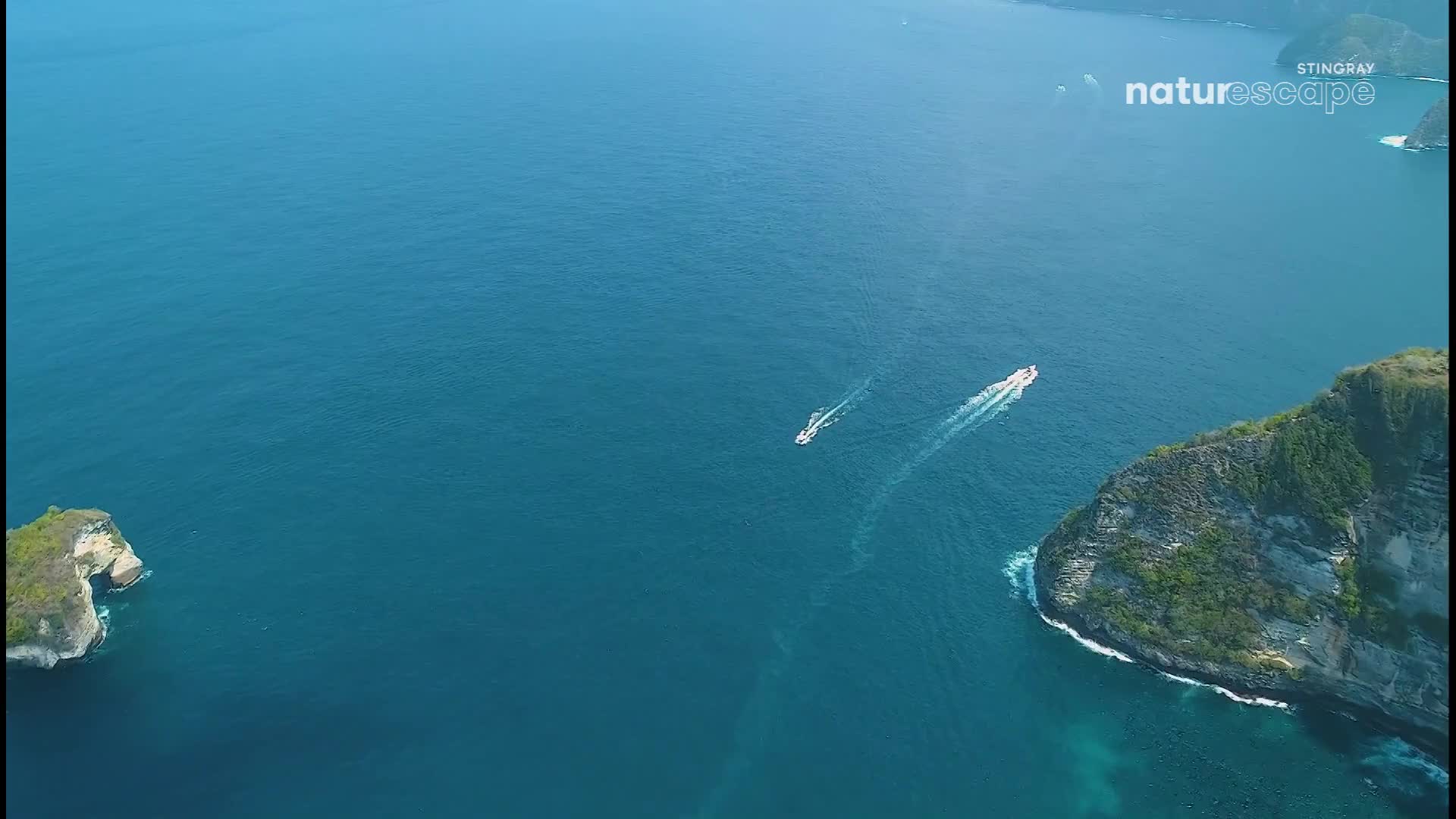 Two white boats are cutting through the deep blue water, leaving foamy trails behind them. A small, rocky island and a larger, green-covered cliff border the scene.
