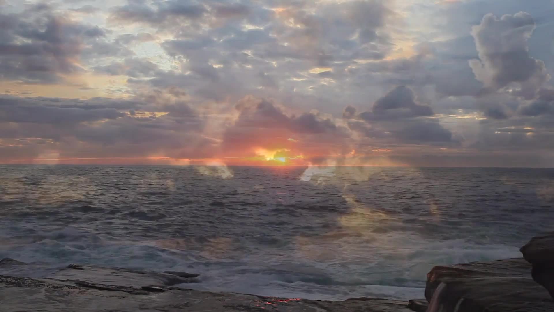 The sun is setting over the ocean, its light breaking through the clouds. Waves crash against the rocky shore, creating a foamy white edge.
