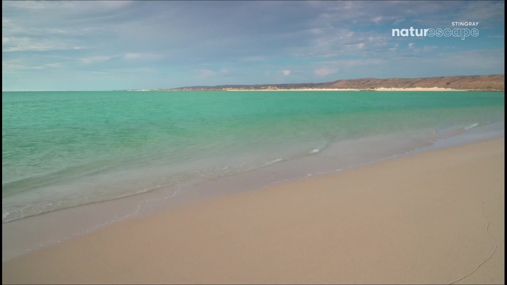 Gentle waves lap the shore, their foamy edges dissolving into the soft sand. The turquoise water stretches out to meet a distant, low-lying coastline under a cloudy sky.
