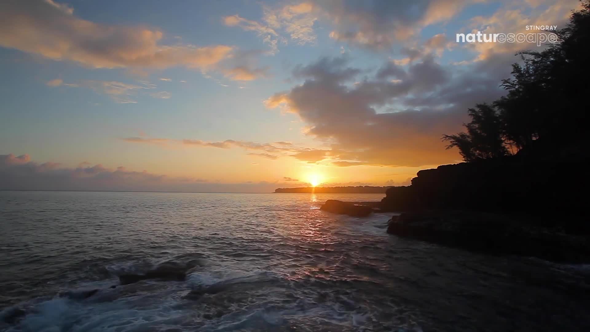The sun dips below the horizon, painting the sky with warm hues as the waves gently lap against the rocky shore. Dark silhouettes of trees and cliffs frame the scene, their edges softened by the fading light.
