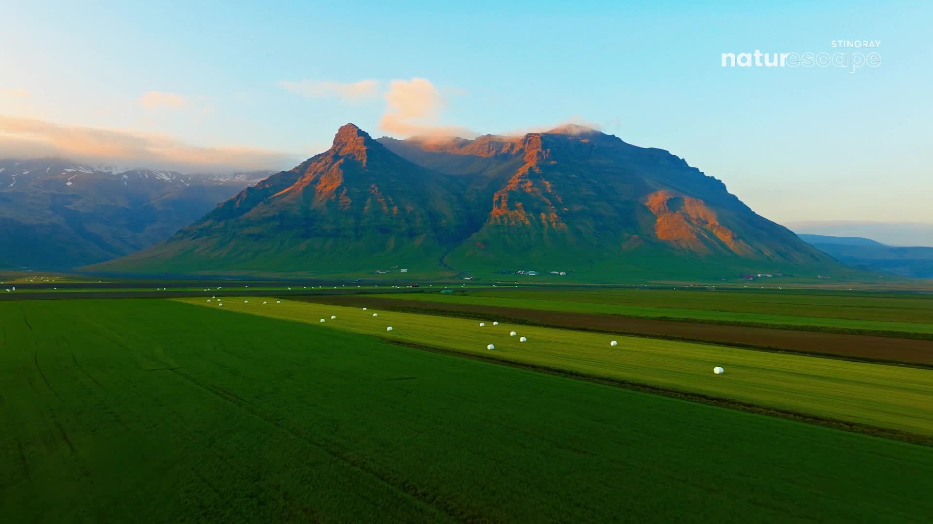 The sun catches the rugged peaks of the mountains, casting long shadows across the vibrant green fields. Round hay bales dot the landscape, waiting to be gathered.