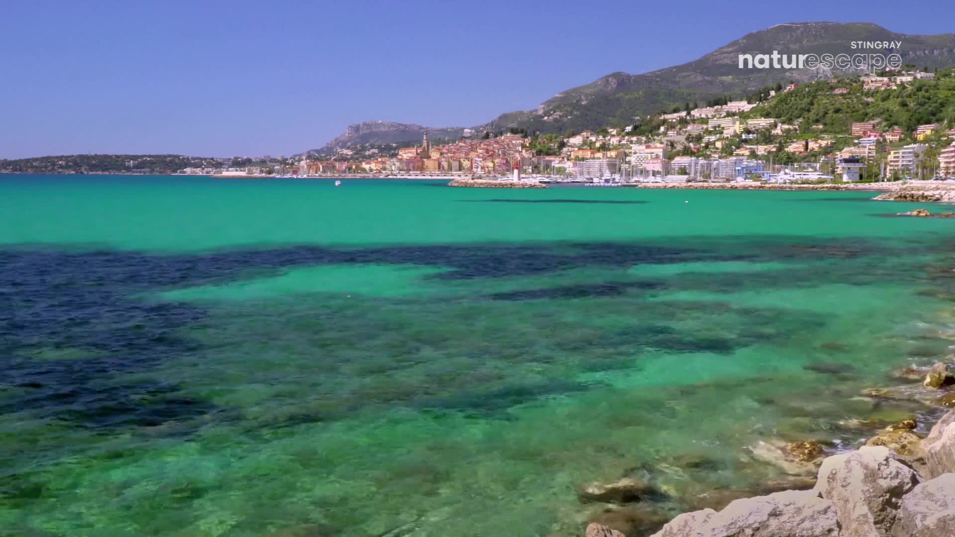 The clear turquoise water laps gently against the rocky shore. A picturesque town with terracotta roofs climbs the hillside overlooking the bay.
