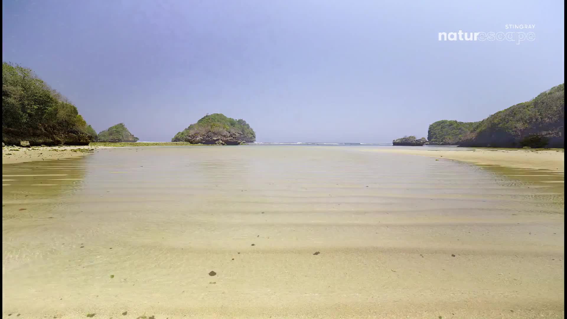 The shallow water reflects the clear sky, revealing the rippled sand beneath. Rocky, tree-covered islets rise from the calm sea.