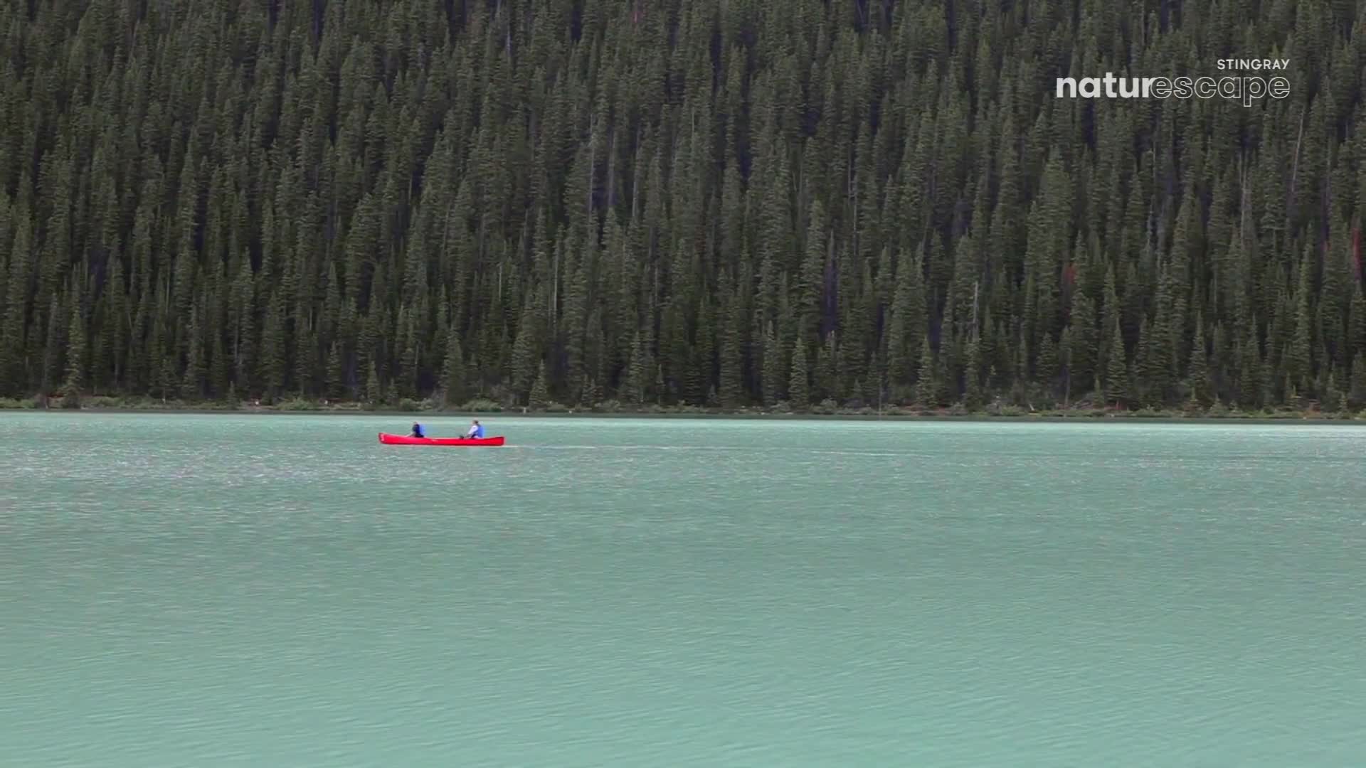 A bright red canoe glides across the turquoise water. Two people paddle gently, their figures small against the backdrop of a dense, dark green forest.