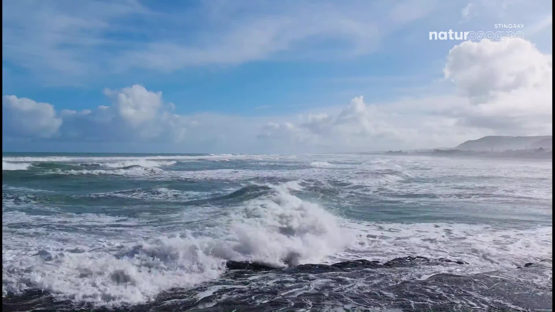 Waves crash against dark rocks, sending white spray into the air. The ocean stretches out under a sky of blue and white clouds.