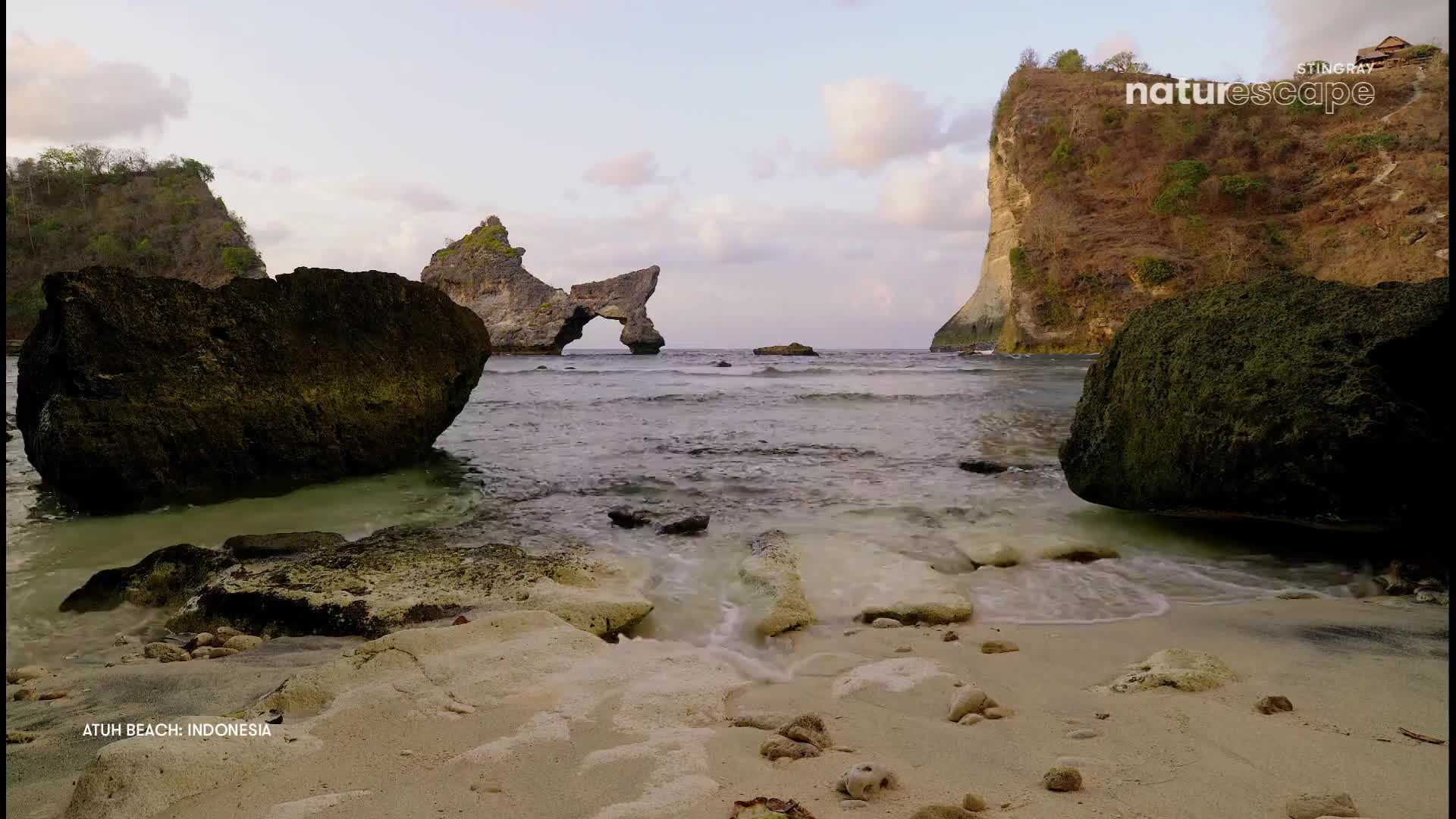 Waves gently wash over the sandy shore, revealing scattered rocks. In the distance, a natural archway stands against the ocean's horizon.