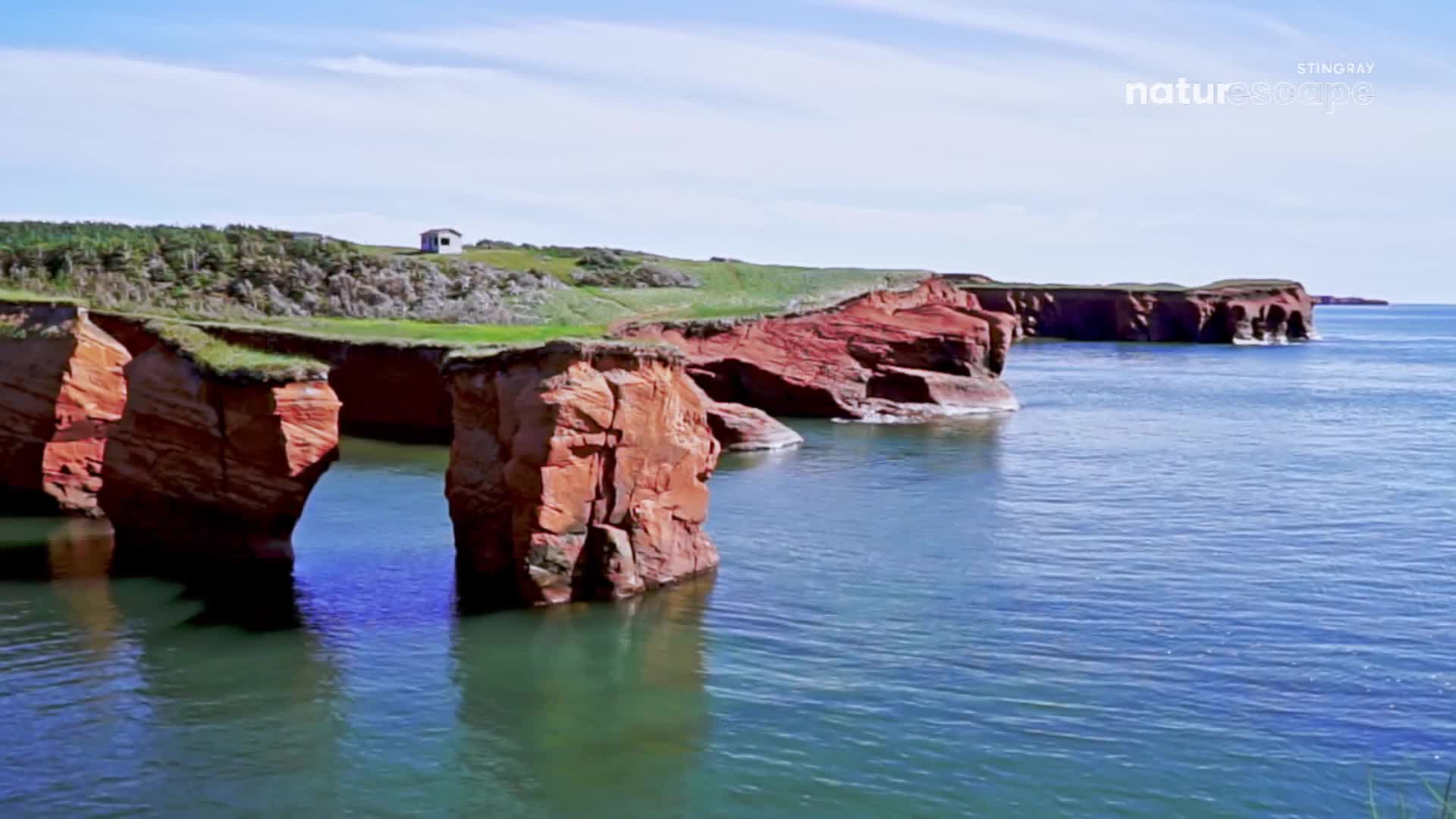 The red sandstone cliffs of Prince Edward Island rise from the calm, blue water. A small white building sits atop a grassy hill overlooking the coastline.