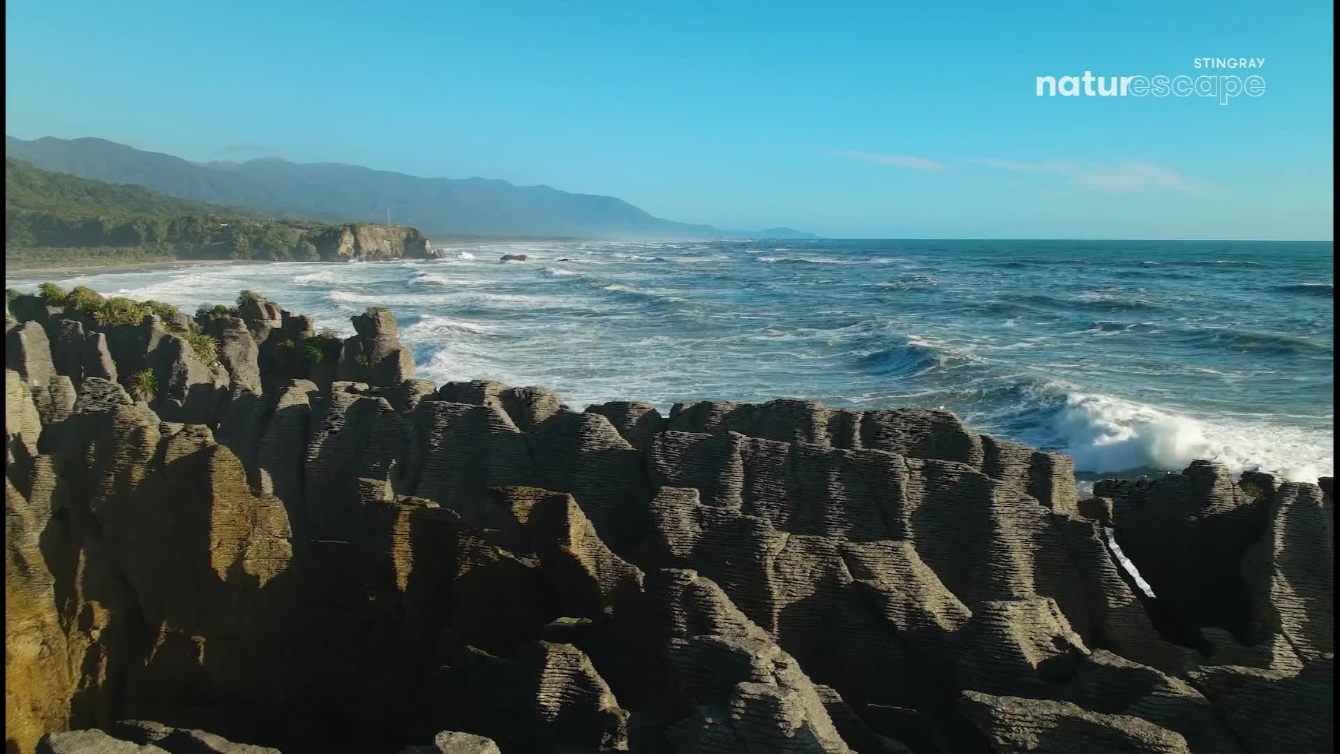 Waves crash against the Pancake Rocks, their white foam contrasting with the dark, stacked stone. The ocean stretches out to a hazy horizon under a clear blue sky.