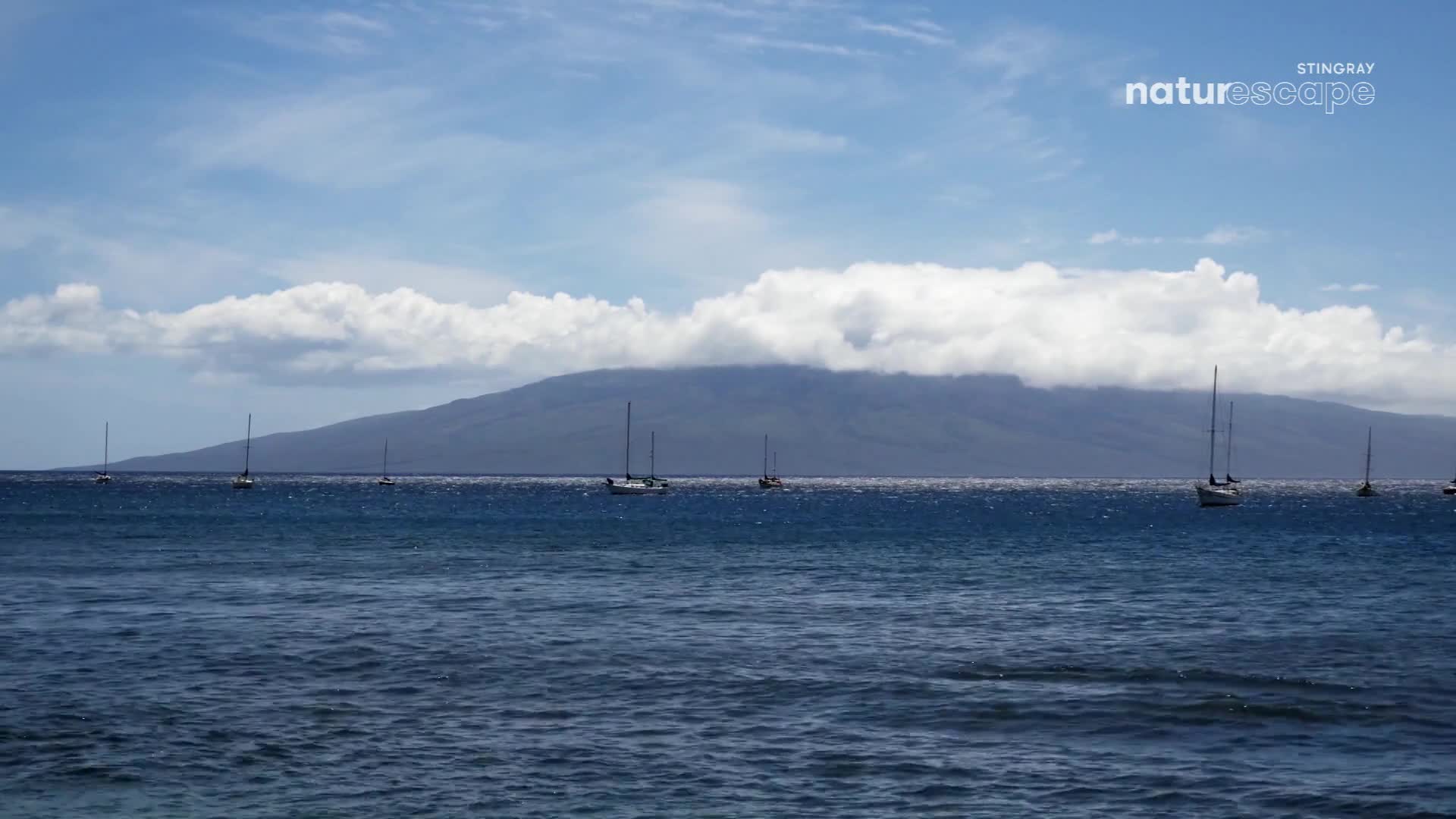 Several sailboats drift on the deep blue water. A large, cloud-covered mountain looms in the distance.