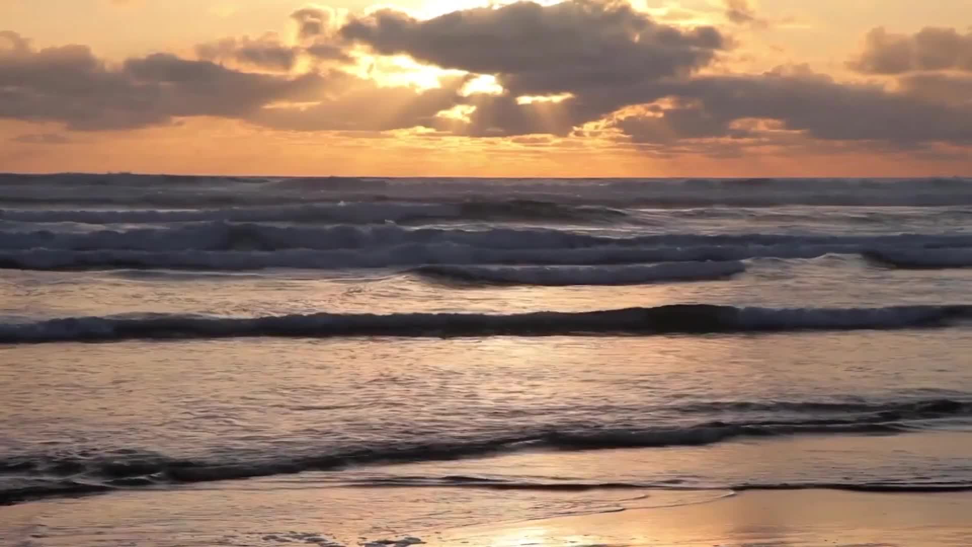 Sunlight breaks through the clouds over the Pacific Ocean, casting a warm glow on the incoming waves. The water rolls in, each crest a darker line against the lighter foam.