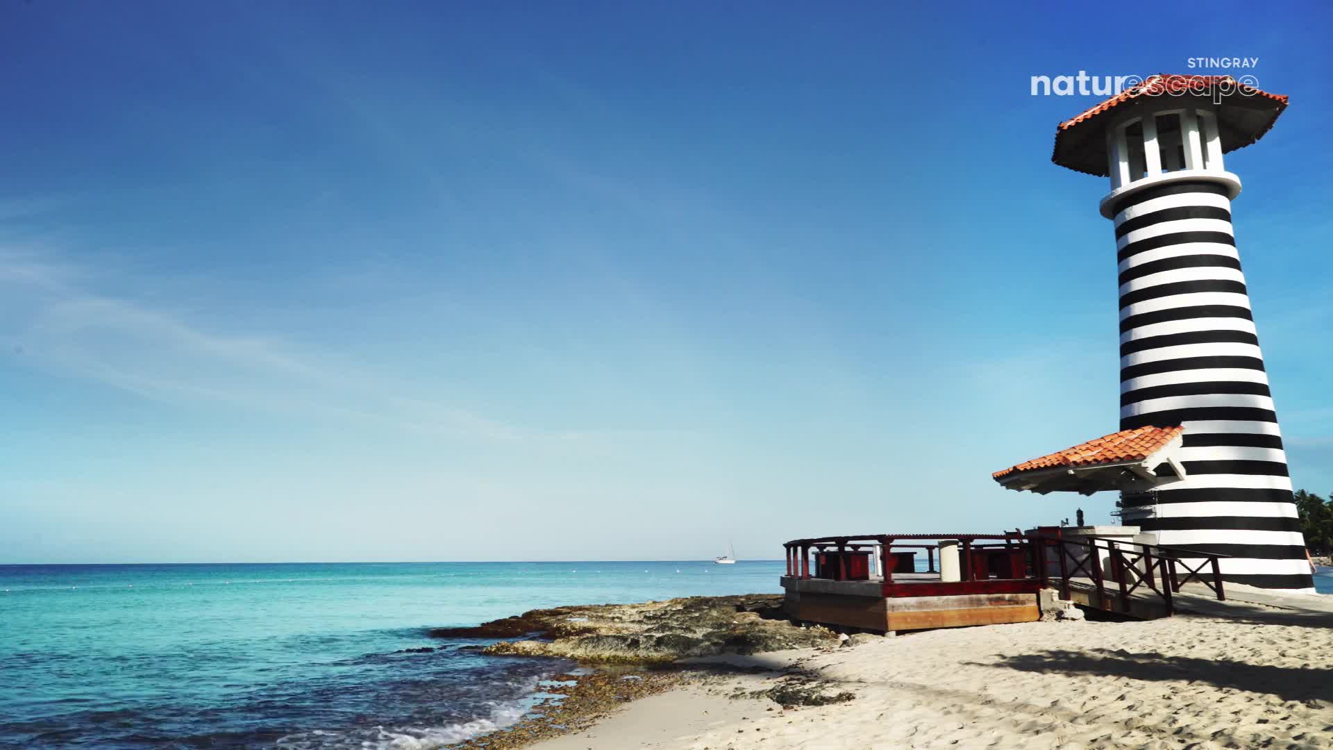 The turquoise water laps gently onto the sandy shore, a lone sailboat glides across the horizon. A striking black and white striped lighthouse stands tall beside a wooden deck, its red-tiled roof gleaming under the bright sun.