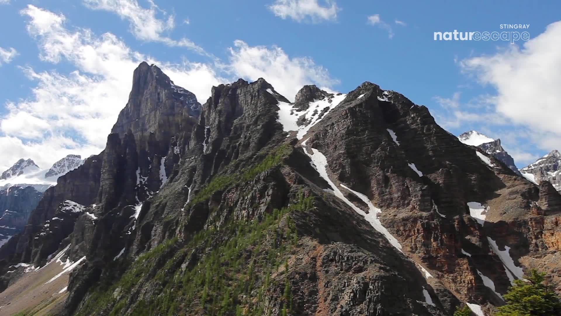 Jagged peaks rise sharply against a bright blue sky, their dark rock faces streaked with lingering snow. Patches of green pine trees cling to the steep slopes, a testament to the resilience of life in this Canadian landscape. Jagged peaks rise sharply against a bright blue sky, their dark rock faces streaked with lingering snow. Patches of green pine trees cling to the steep slopes, a testament to the resilience of life in this Canadian landscape.