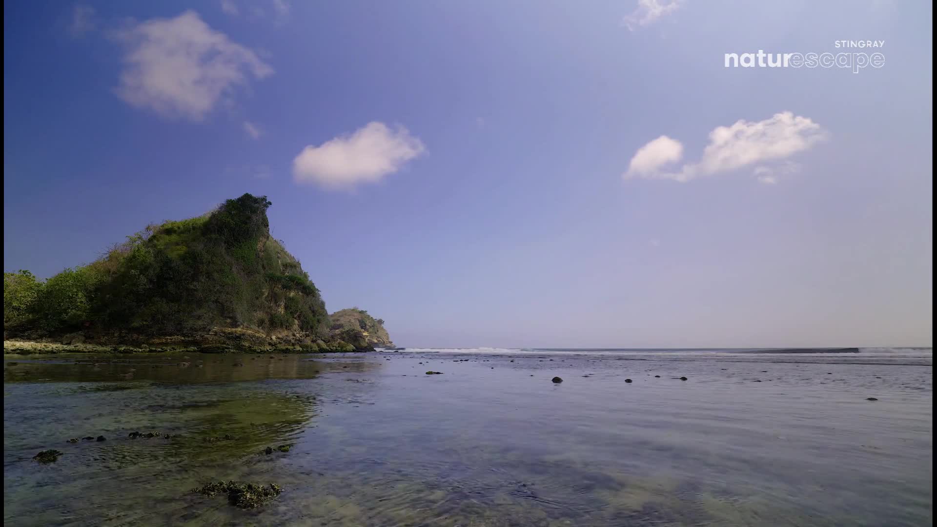 The tide is out, revealing a rocky shoreline and the clear, shallow water of the ocean. A lush, green cliff rises from the left, its peak silhouetted against the bright blue sky dotted with white clouds. The tide is out, revealing a rocky shoreline and the clear, shallow water of the ocean. A lush, green cliff rises from the left, its peak silhouetted against the bright blue sky dotted with white clouds.