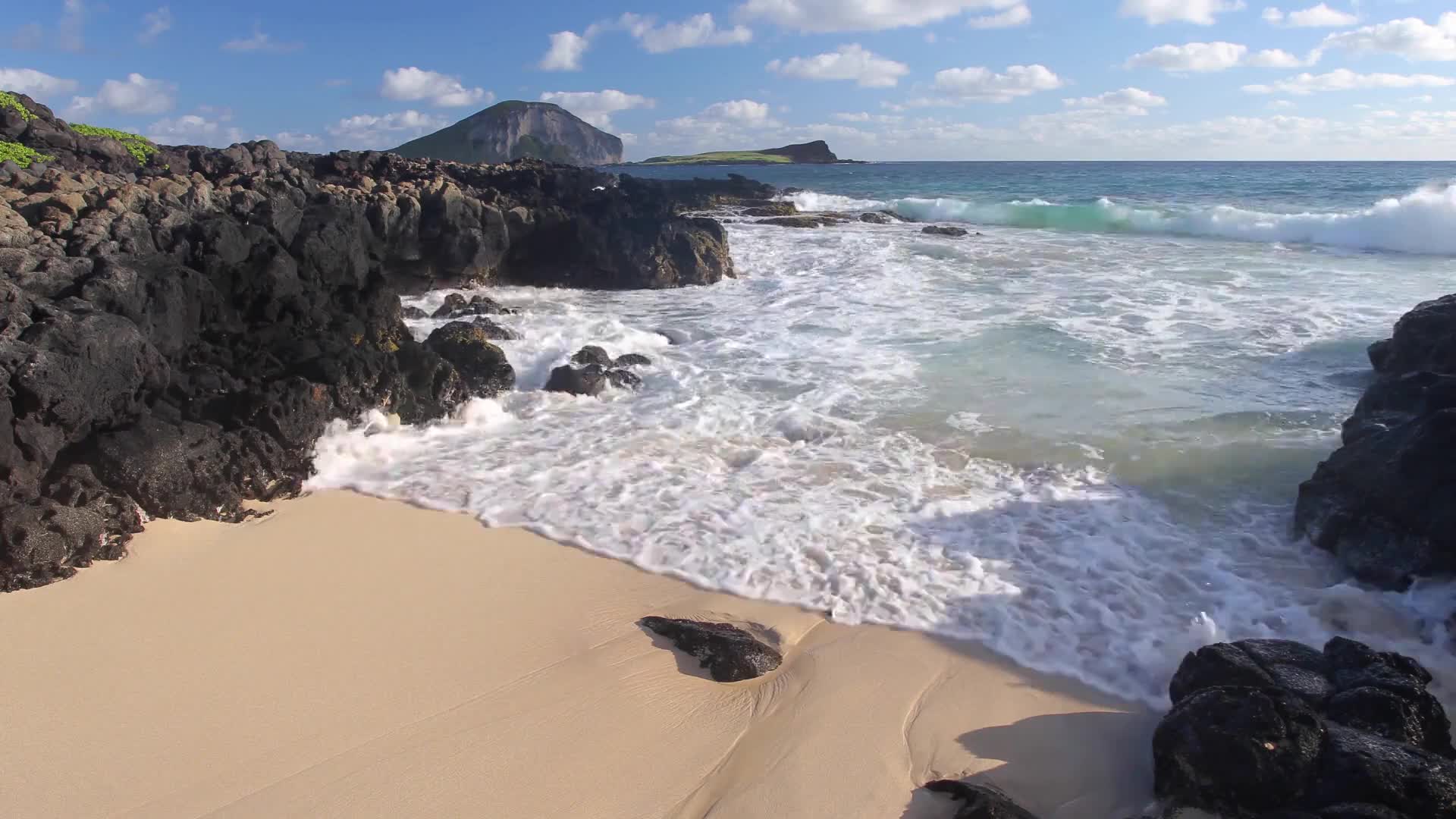 Waves crash against the dark, jagged rocks, sending white foam onto the pale sand. In the distance, two islands rise from the turquoise ocean under a bright blue sky.