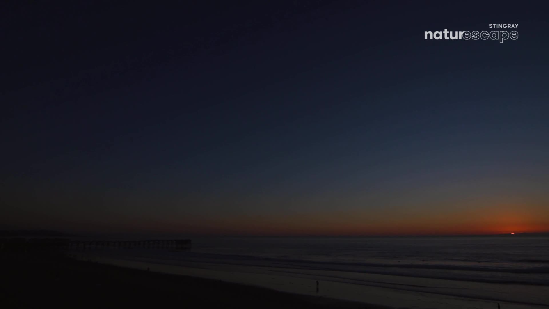The sun dips below the horizon, painting the sky with fiery orange and deep blue. Waves gently roll onto the sandy shore, leading to a distant pier.