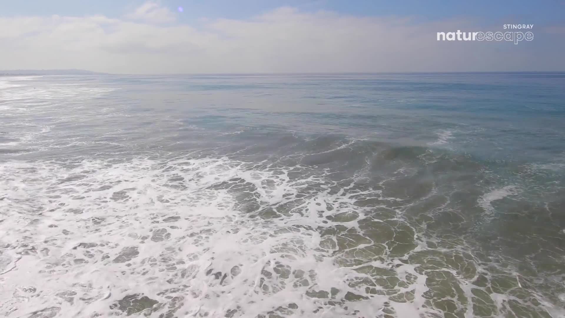 Waves are rolling in, their white foam spreading across the green water. The sky above is a hazy blue with scattered clouds.